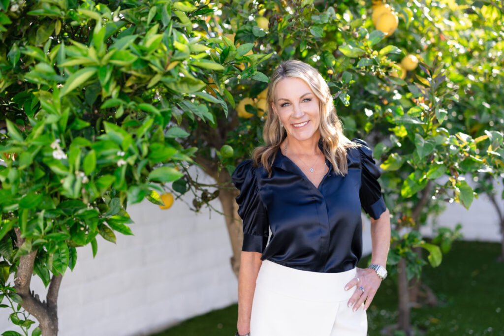 Smiling woman in a black blouse and white pants stands by a vibrant lemon tree. The setting is bright, with a cheerful and relaxed atmosphere.