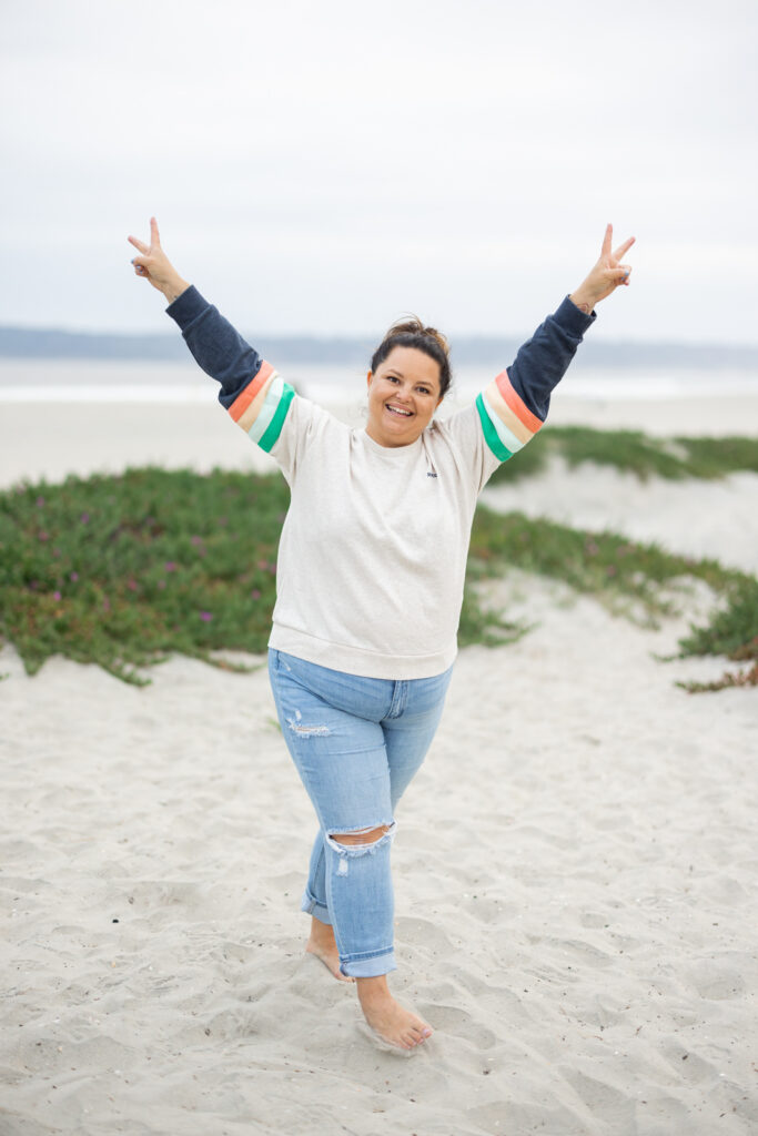 A woman joyfully raises both arms in a "peace" sign on a sandy beach. She's wearing a colorful sweater and ripped jeans, with green dunes behind her. | Meg Marie Photography