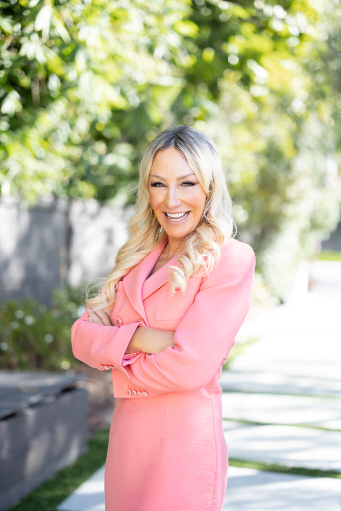 A woman with long blonde hair smiles warmly, wearing a pink blazer, standing outdoors with arms crossed. Sunlit greenery surrounds her, creating a cheerful, vibrant atmosphere. | Meg Marie Photography