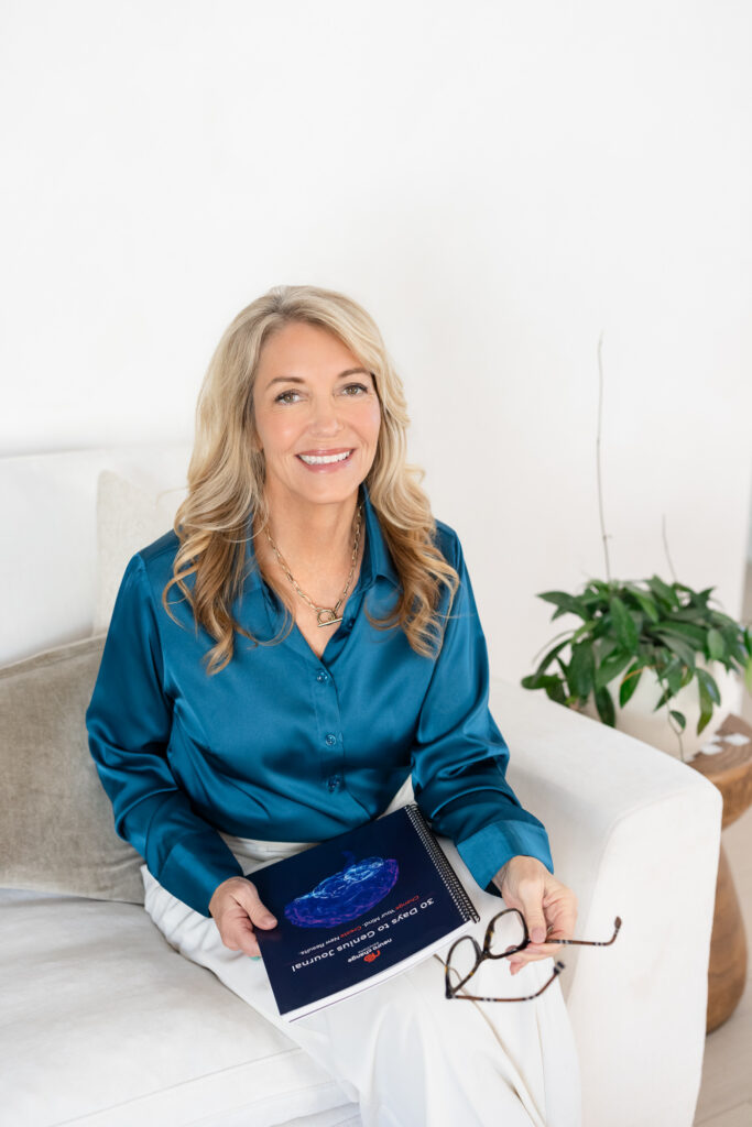 Smiling woman in a teal blouse sits on a white couch, holding eyeglasses and a booklet. Plant in the background. Bright, professional atmosphere. | Meg Marie Photography