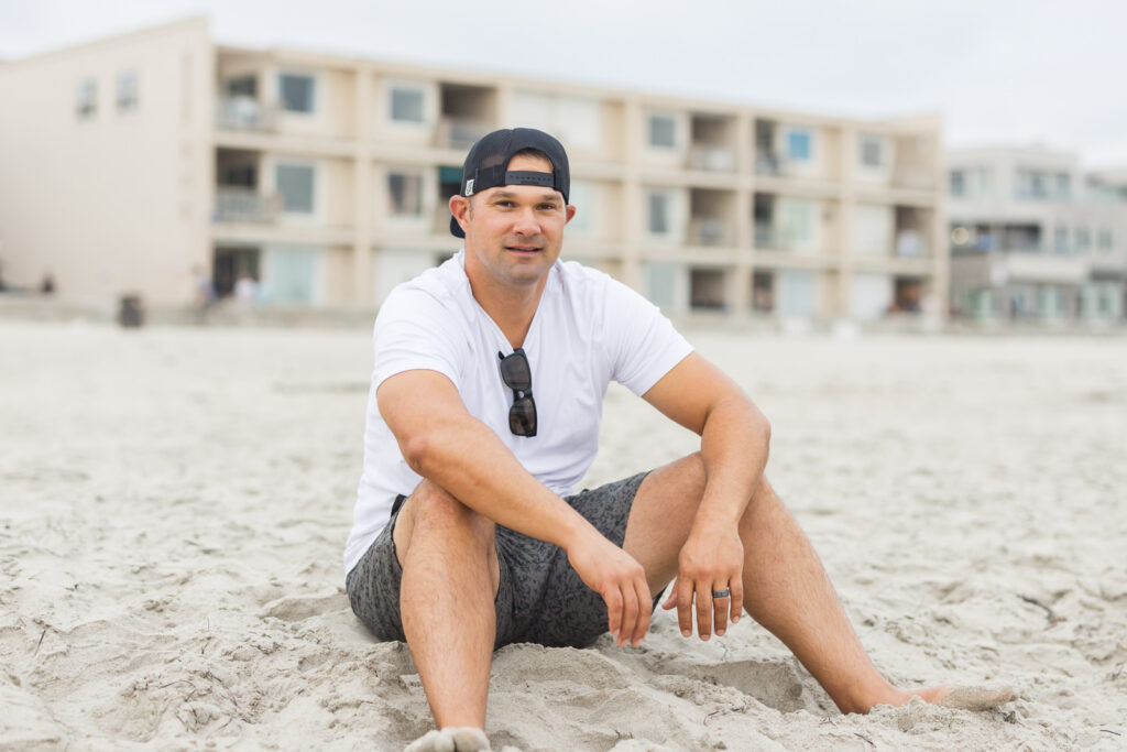 A smiling man in a white t-shirt and cap sits on a sandy beach. Sunglasses hang from his shirt. Residential buildings are blurred in the background. | Meg Marie Photography