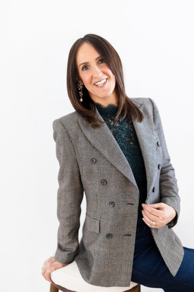 Smiling woman with brown hair, wearing a gray plaid blazer and a dark lace top, sits against a white background, conveying a confident and approachable tone. | Meg Marie Photography