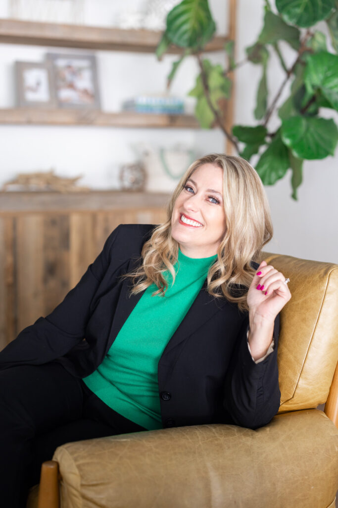 Smiling person in a black suit and green top, sitting on a yellow chair. Background shows wooden shelves with decor and a leafy plant. | Meg Marie Photography