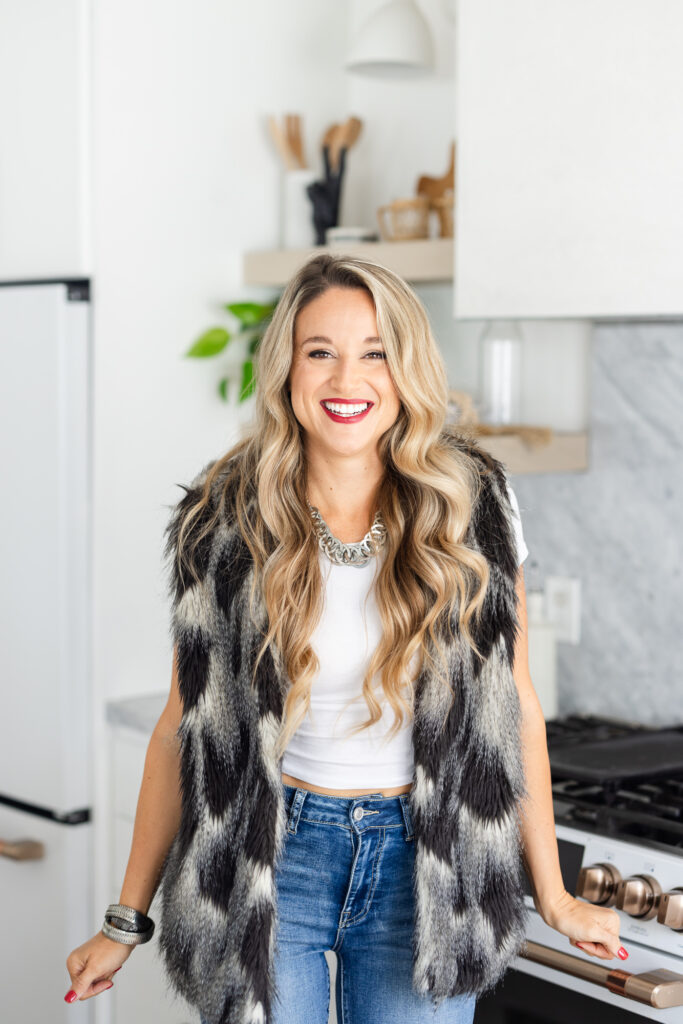 Smiling person with long blonde hair in a white shirt and patterned fur vest, standing in a modern kitchen. Bright, cheerful atmosphere. | Meg Marie Photography