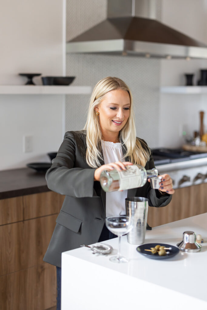 Woman with long blonde hair in a blazer, cheerfully pours a drink into a cocktail shaker in a modern kitchen. Martini glass and olives nearby. | Meg Marie Photography