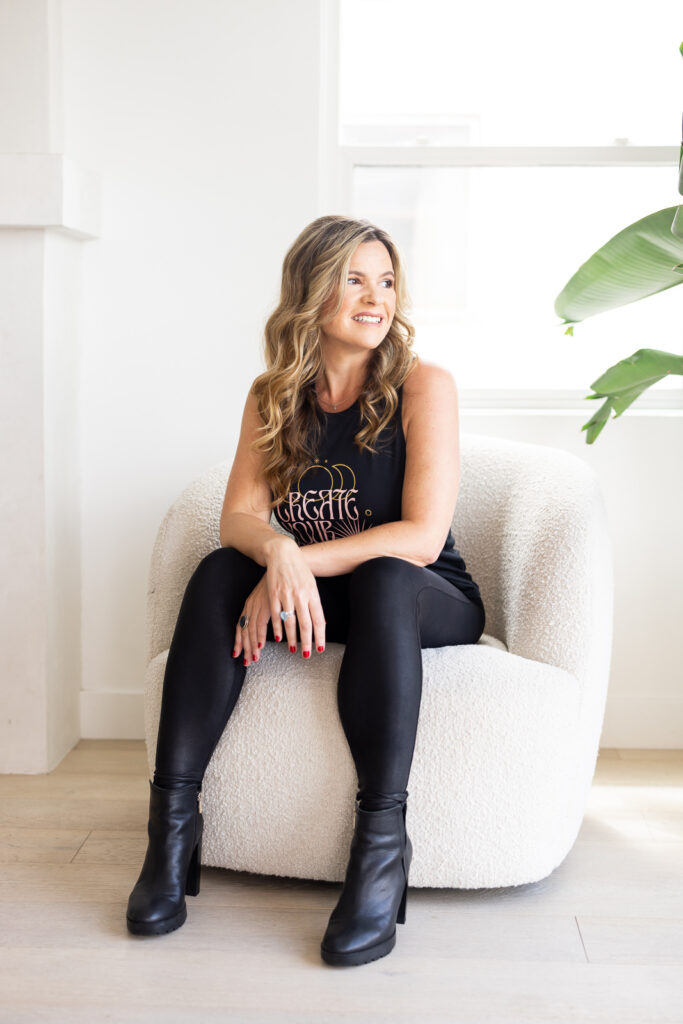 Woman with long hair, smiling, seated on a white fuzzy chair. Wearing a black outfit and boots, in a bright room with a plant in the background. | Meg Marie Photography