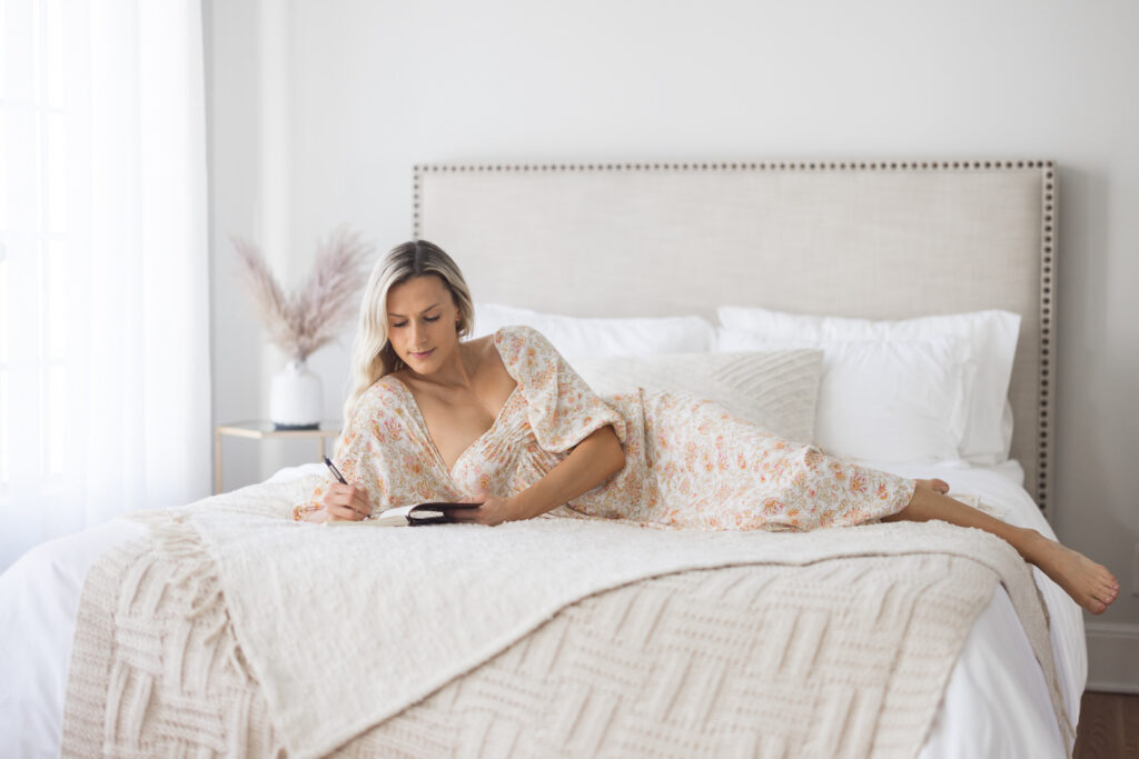 Woman in floral dress lies on a bed with white linens, writing in a journal. Soft light filters through a window, creating a serene and peaceful ambiance. | Meg Marie Photography