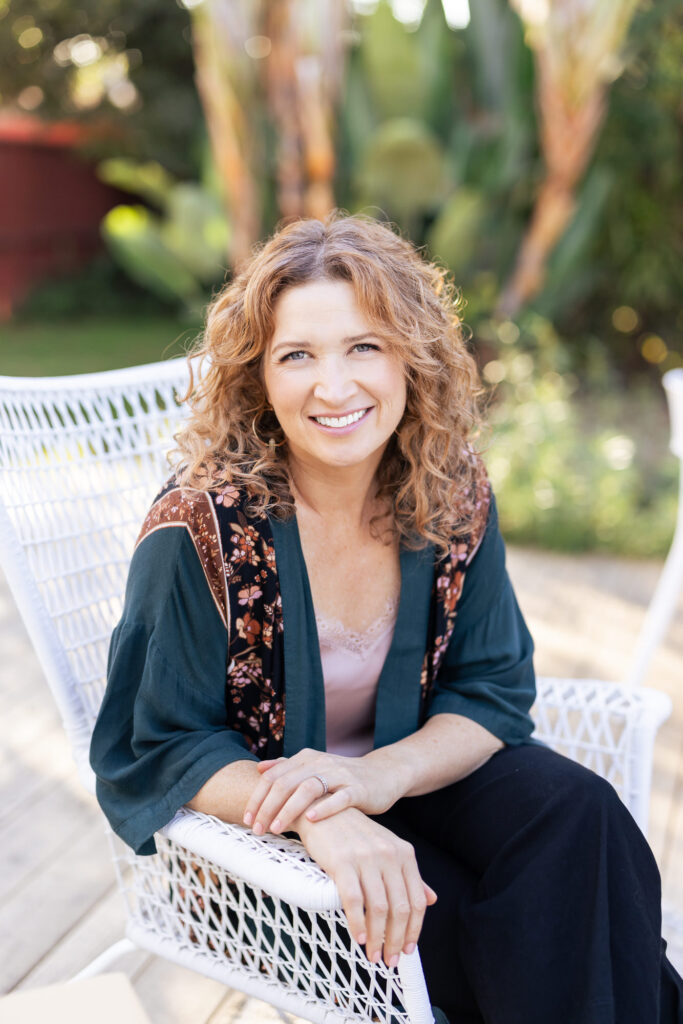 Smiling woman with curly hair sits on a white wicker chair in a sunlit garden. She's wearing a floral-patterned shawl and a pink top, exuding warmth. | Meg Marie Photography