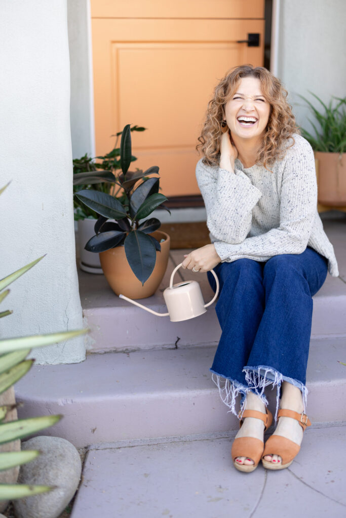 A woman with curly hair sits on pink steps, laughing joyfully. She holds a watering can next to a potted plant. The scene is bright and cheerful. | Meg Marie Photography