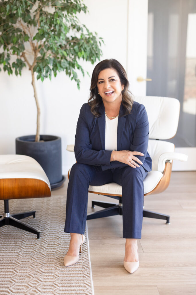 A woman in a navy suit sits on a white chair, smiling warmly. She is in a modern office with a large potted plant and wooden floor. Professional and welcoming. | Meg Marie Photography