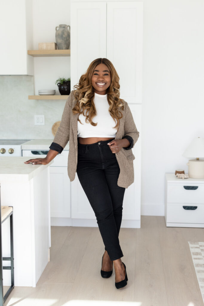 Smiling woman with long hair stands in a bright kitchen. She wears a white top, black pants, and a cardigan, conveying warmth and confidence. | Meg Marie Photography