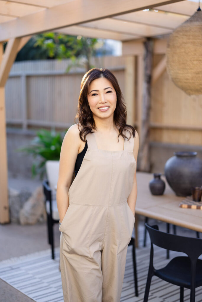 A smiling woman in a beige jumpsuit stands outdoors in a modern patio. She exudes warmth and confidence, surrounded by a wooden table and greenery. | Meg Marie Photography