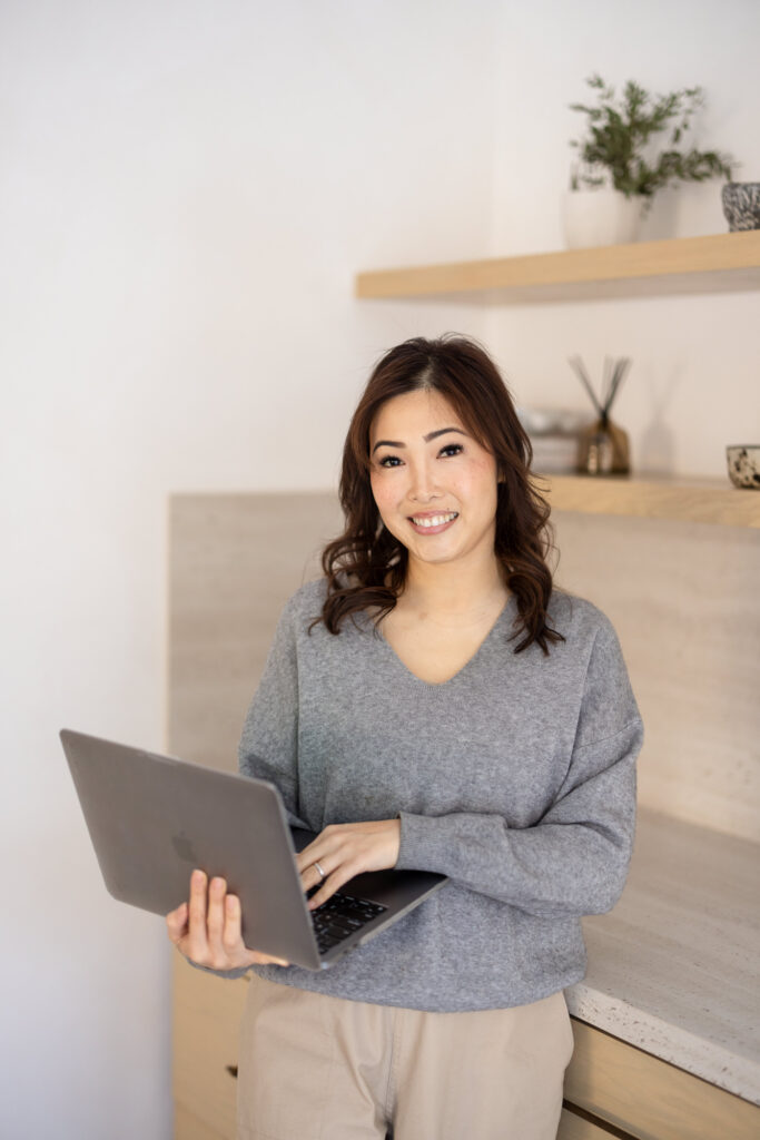 Smiling woman with wavy hair in a gray sweater holds a laptop in a modern kitchen. Shelves with plants and decor create a cozy, inviting atmosphere. | Meg Marie Photography