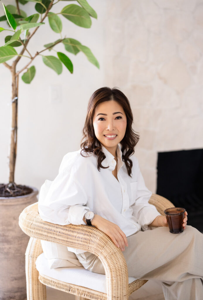 A woman in a white blouse sits in a wicker chair, smiling and holding a cup. A large green plant is in the background, creating a calm, inviting atmosphere. | Meg Marie Photography