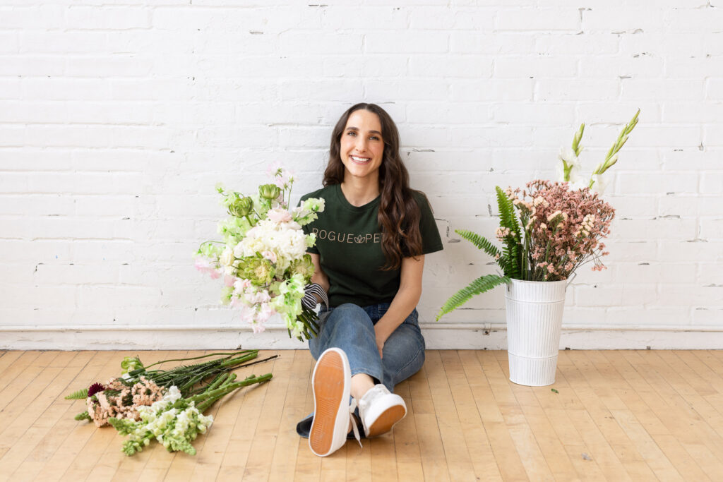 Woman sitting on wooden floor, smiling and holding a bouquet of flowers. She wears a green T-shirt and jeans. Buckets of flowers are on either side. | Meg Marie Photography