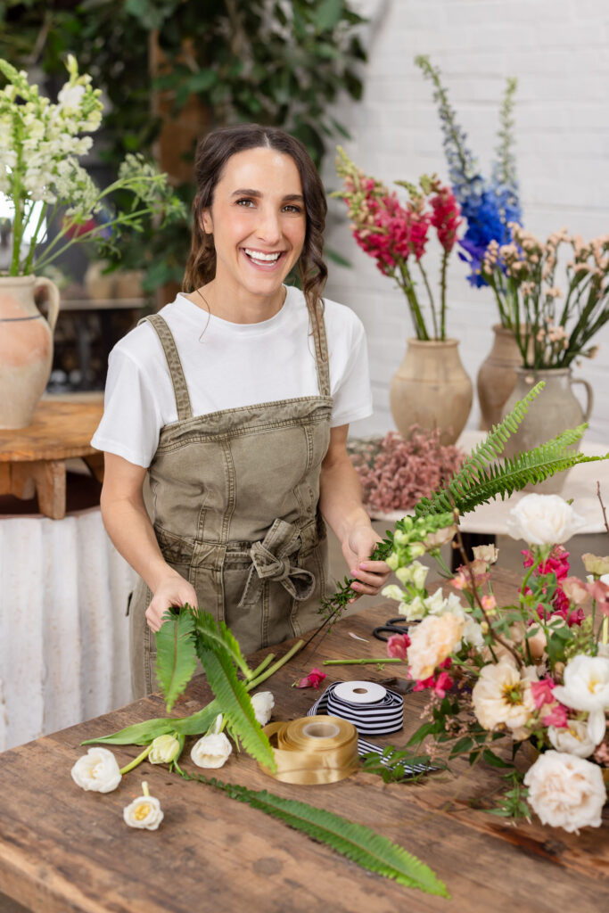 A smiling woman in a white shirt and olive overalls arranges flowers on a wooden table. Colorful blooms and vases are in the background. | Meg Marie Photography