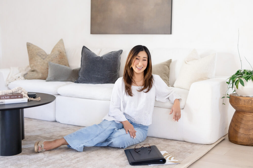 A smiling woman sits on the floor in a cozy living room, leaning against a white sofa with plush pillows. A coffee table and plant are nearby. | Meg Marie Photography