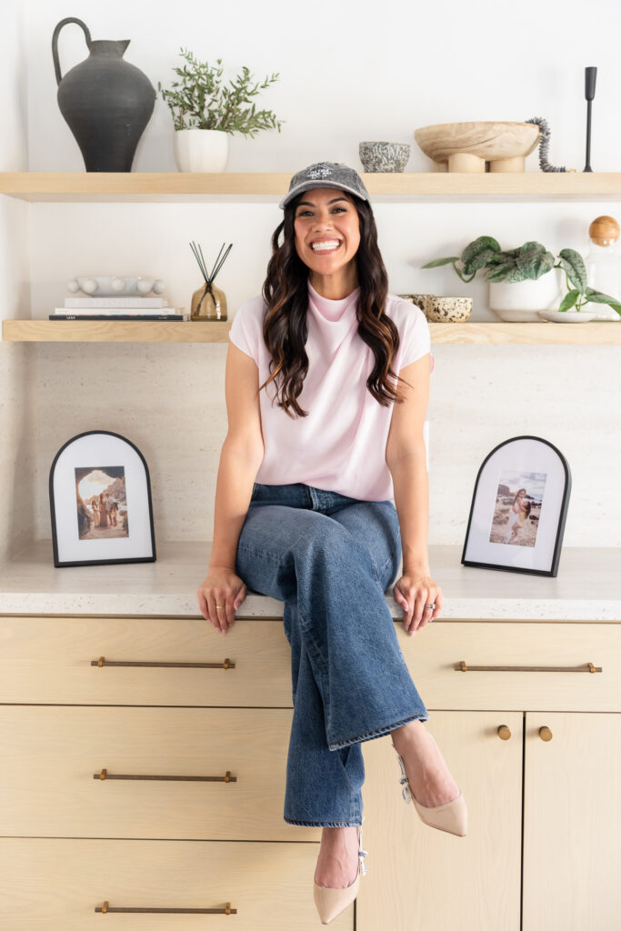 A woman in a pink shirt and jeans sits on a kitchen counter, smiling warmly. The kitchen is decorated with plants, pottery, and framed photos, creating a cozy atmosphere. | Meg Marie Photography