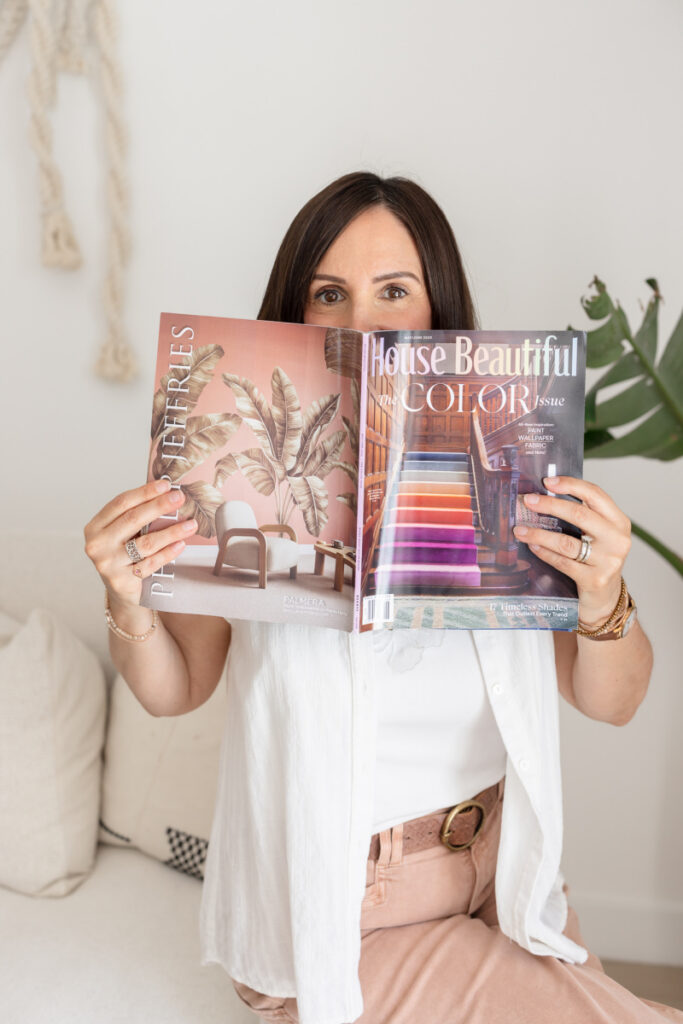 A woman in a light outfit holds up two decorative interior magazines, partially covering her face. Behind her, a plant and a minimalist room setting. | Meg Marie Photography