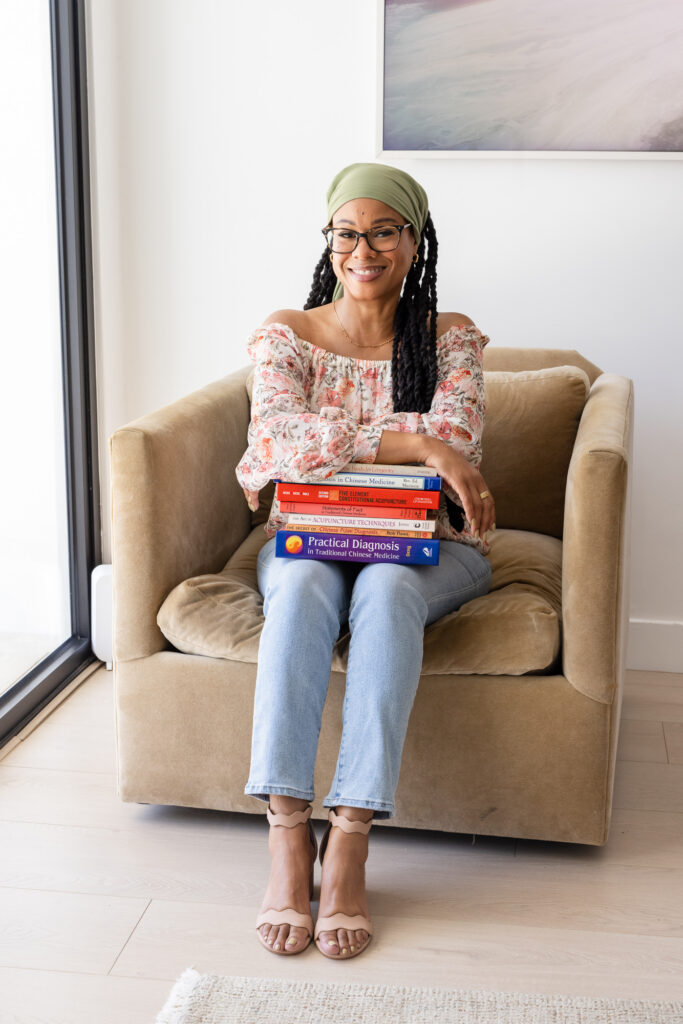 A smiling woman sits in a beige armchair holding a stack of colorful books, including one titled *Practical Diagnosis in Traditional Chinese Medicine*. She wears a floral off-shoulder top, jeans, a green head wrap, and glasses. The bright, airy room has white walls and natural light. | Meg Marie Photography