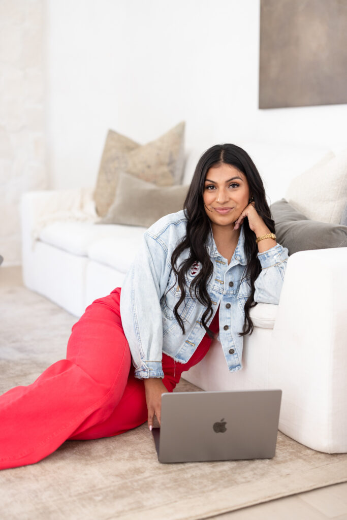A young woman with long dark hair reclines on a beige carpet leaning against a white sofa, wearing a light denim jacket and red wide-leg pants, with an open silver MacBook laptop in front of her. The bright, airy room features neutral tones and soft natural lighting. | Meg Marie Photography