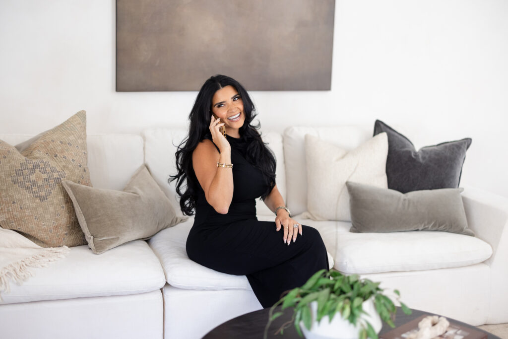 Smiling woman with long black hair sitting on a white sectional sofa while talking on a smartphone, wearing a fitted black sleeveless dress and gold jewelry. Neutral-toned decorative pillows, a minimalist wall art piece, and a modern coffee table with greenery create an elegant, contemporary living room setting. Bright natural lighting and soft beige accents give the image a polished lifestyle and personal branding aesthetic.