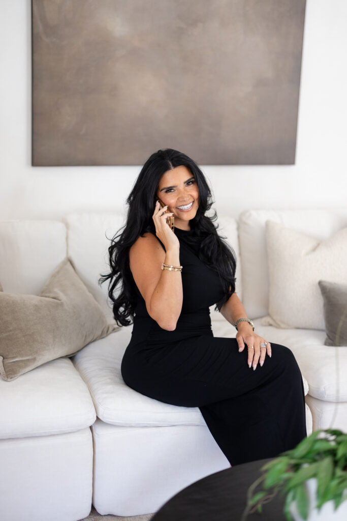 Woman with long dark hair in a black outfit sits on a white sofa, smiling while talking on a gold phone. Bright, neutral-toned living room with abstract wall art behind her. | Meg Marie Photography