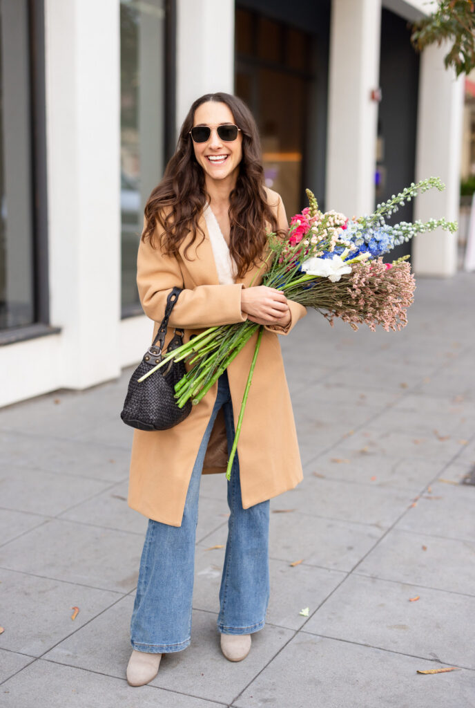 A smiling woman with long wavy brown hair holds a large colorful wildflower bouquet on a city sidewalk. She wears a camel coat, wide-leg jeans, tan ankle boots, and dark sunglasses, with a woven black bag over her shoulder. | Meg Marie Photography