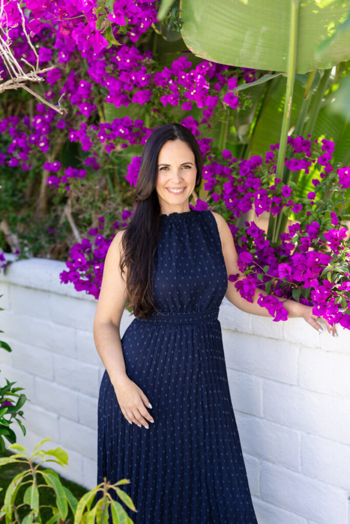 A woman with long dark hair stands smiling in a navy pleated midi dress against a white brick wall, surrounded by vibrant magenta bougainvillea blooms and lush green tropical foliage in bright natural light. | Meg Marie Photography