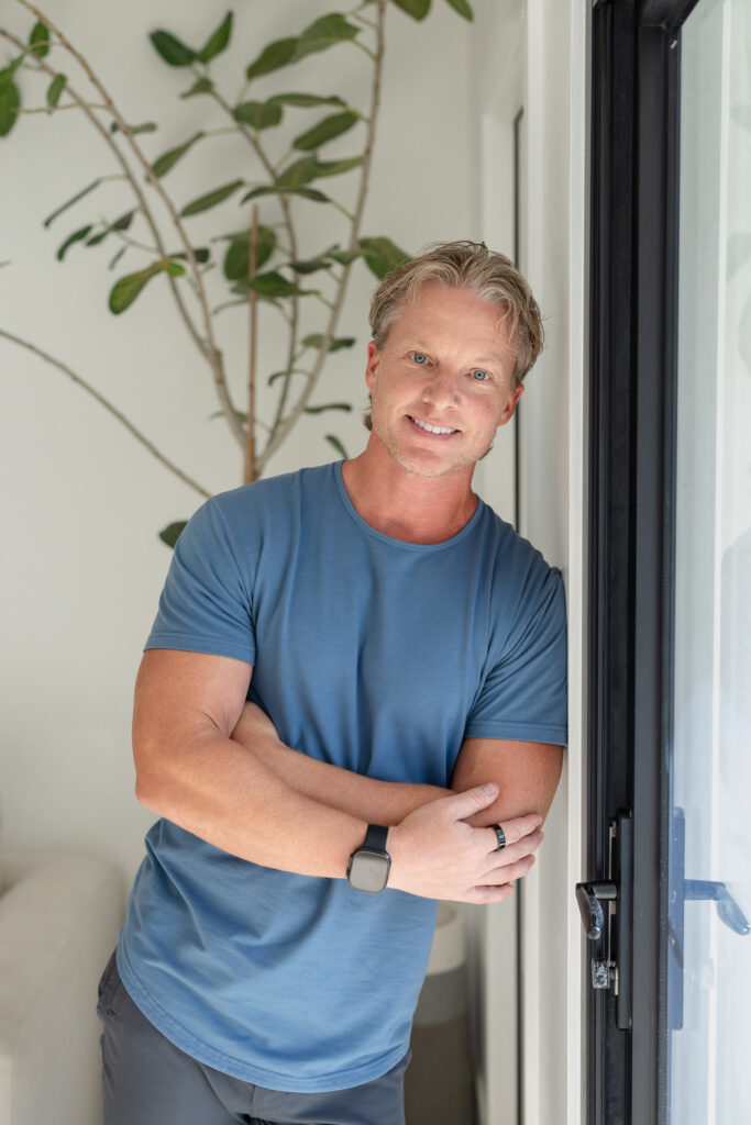 A smiling man with blond hair leans against a dark door frame with arms crossed, wearing a blue t-shirt and a smartwatch. Bright, airy indoor setting with a green plant in the background. | Meg Marie Photography