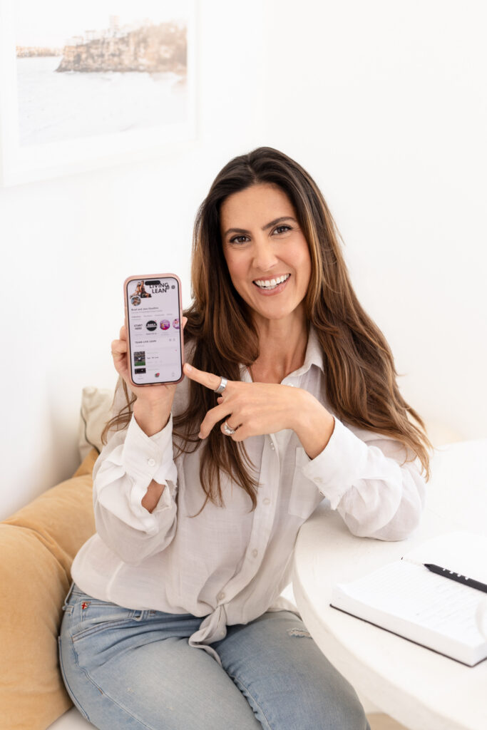 A smiling woman with long brown hair sits on a beige sofa, pointing at a smartphone she holds up. She wears a white linen shirt and jeans. The bright, minimal room has soft natural lighting. | Meg Marie Photography