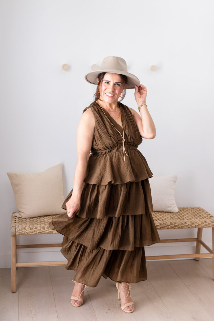 A woman stands smiling in a tiered olive-brown maxi dress with a wide-brim tan hat and nude heeled sandals. She poses in a bright, minimalist room with a woven bench and neutral cushions behind her. | Meg Marie Photography