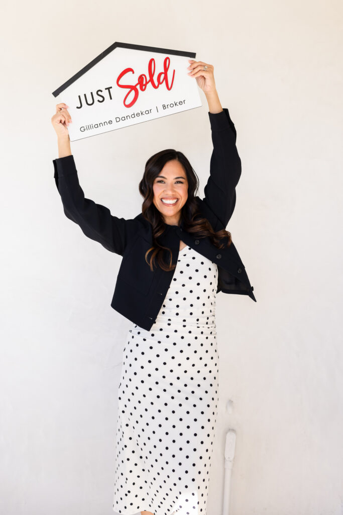 A smiling woman holds a house-shaped "Just Sold" sign overhead with both arms raised. She wears a white polka-dot dress and black jacket against a plain white background. The mood is celebratory and bright. | Meg Marie Photography