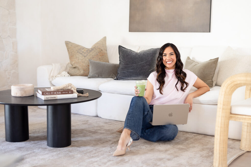 Woman seated on floor, leaning against a sofa with gray pillows, holding a smoothie in one hand and a laptop in the other. Room has a cozy, modern vibe. 