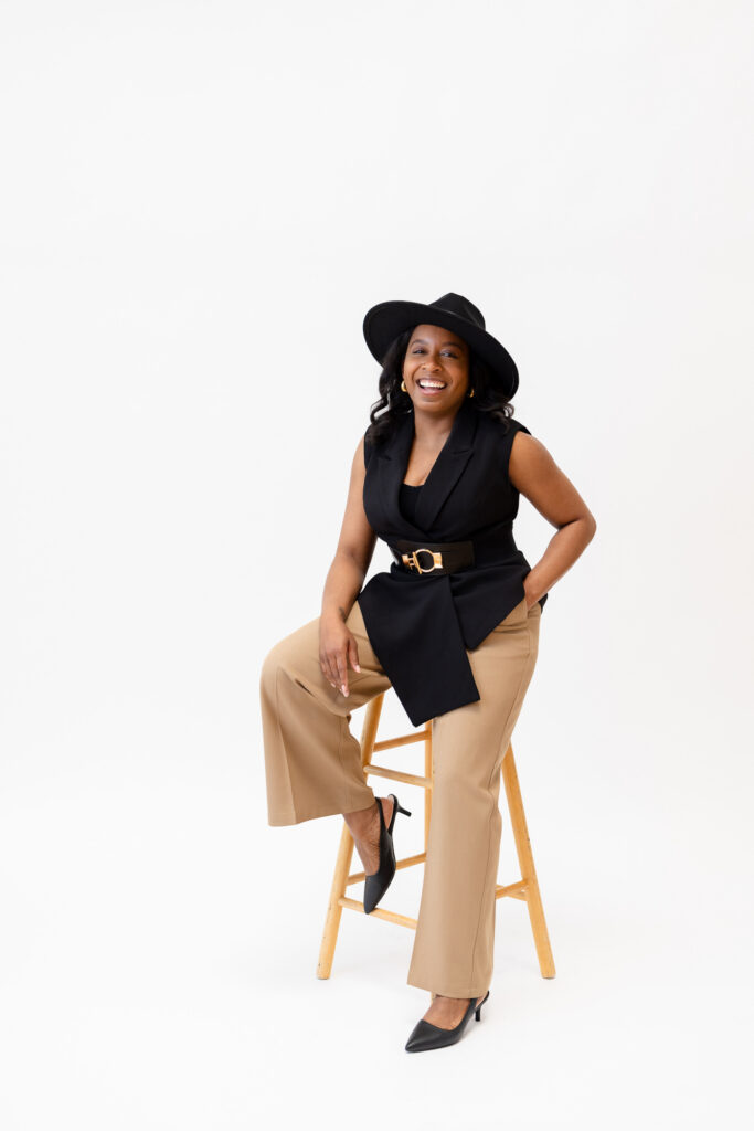 A woman with long dark hair and sunglasses sits laughing in a tall black metal chair, holding a green drink aloft. She wears a gray blazer, dark shorts, and black heels against a plain white background with bright, even lighting. | Meg Marie Photography