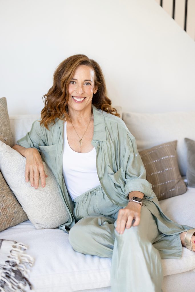 A smiling woman in a sage-green outfit sits comfortably on a white sofa among neutral cushions, creating a relaxed, warm, and welcoming portrait.